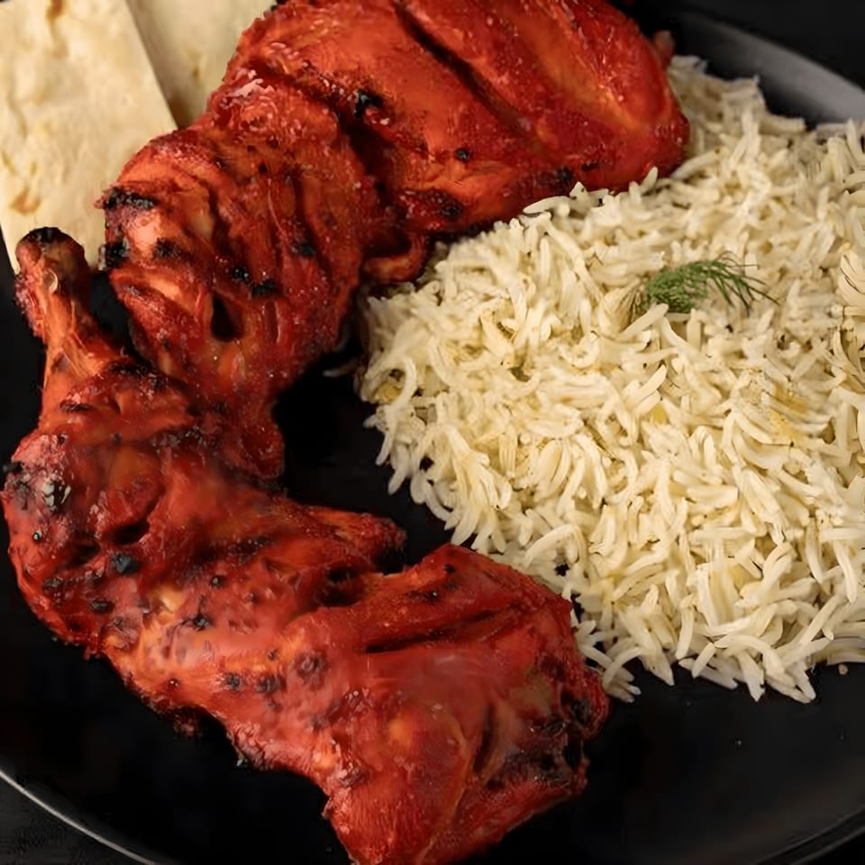 Two pieces of red tandoori chicken with white rice and flatbread on a black plate.