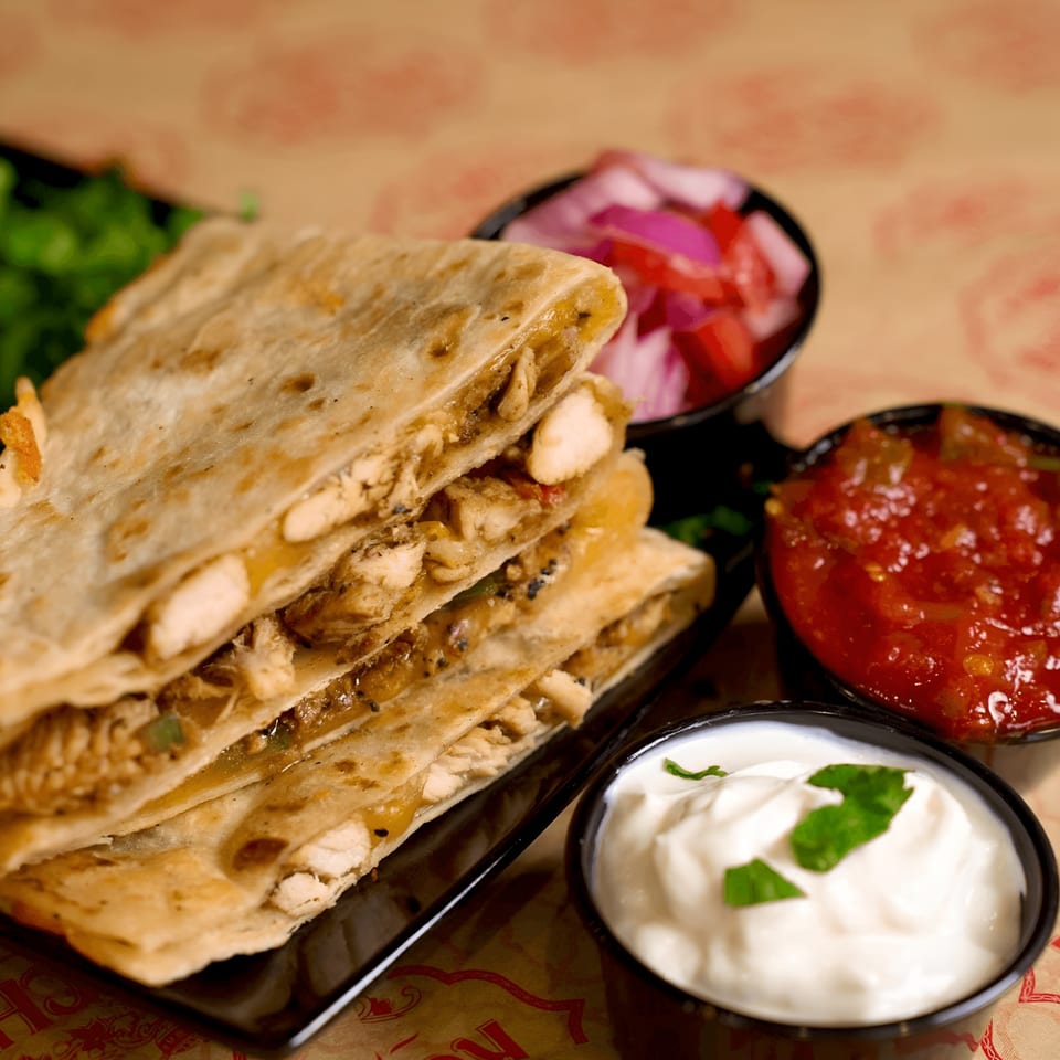 Chicken quesadillas with sides of salsa, sour cream, and pickled red onions on a patterned surface.